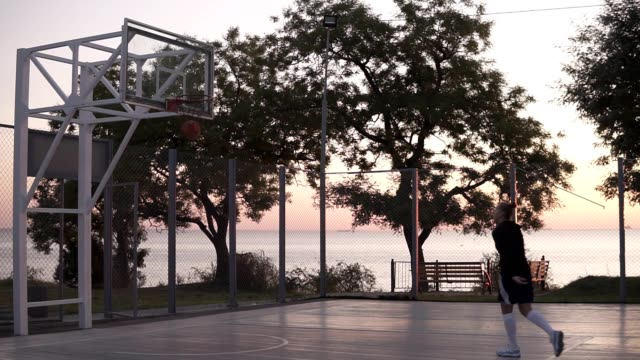 Handhelded-footage-of-young-girl-in-shorts-and-white-golf-socks-make-a-shot-to-the-basketball-net.-Outdoors,-trees-on-the-background.-Morning-dusk