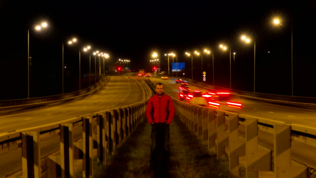 A-man-is-standing-on-the-highway.-Freeway-traffic-blurry-cars-at-night-time-lapse.-Cars-moving-on-road-on-bridge-evening-timelapse.-4K-UHD