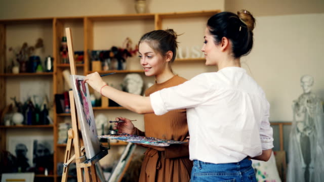 Junge-Frauen-Kunstlehrer-und-Schüler-malen-gemeinsam-sprechen-und-Lächeln-während-des-Unterrichts-im-Kreativstudio.-Bildende-Kunst,-Bildung-und-Menschen-Konzept.