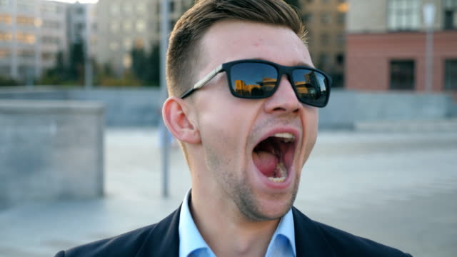 Portrait-of-tired-businessman-in-sunglasses-yawning-outdoor.-Exhausted-sleepy-man-opening-his-mouth-outside.-Angry-guy-yelling-to-camera.-Handsome-male-entrepreneur-screaming-at-city-street.-Close-up-Slow-motion