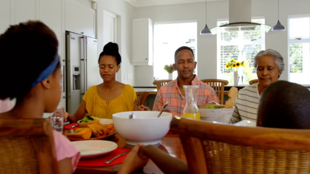 Vista-frontal-de-la-familia-de-generación-negro-multi-orar-juntos-en-mesa-de-comedor-en-hogar-confortable-4k