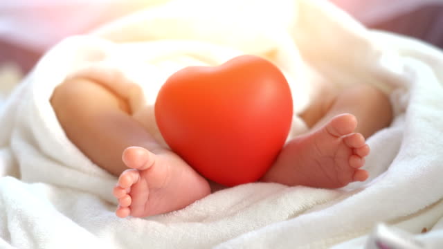 Bebé-recién-nacido-moviendo-el-pie-y-el-corazón-rojo-en-la-luz-hermosa-sobre-fondo-blanco