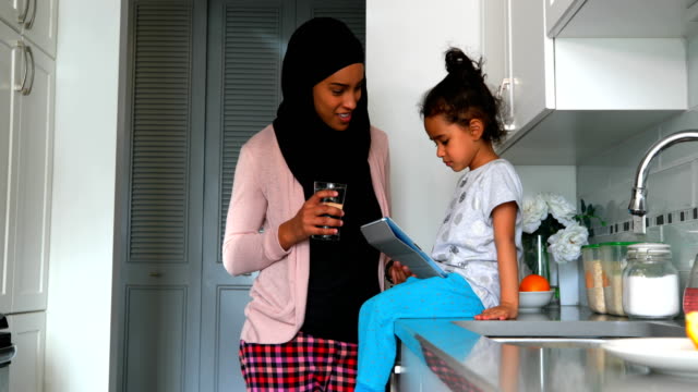 Joven-madre-con-un-hijab-bebiendo-un-vaso-de-agua-cerca-de-su-hija-en-la-cocina-4k