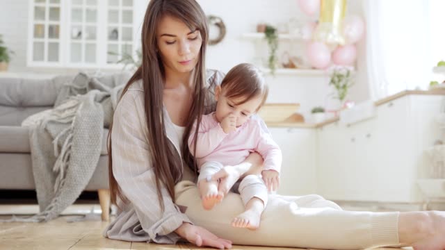 Beautiful-young-mother-sitting-on-floor-at-home-and-playing-with-baby-daughter-on-lap-patting-gently-her-tiny-feet
