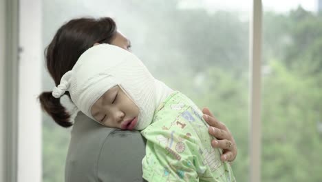 Asiatische-Mutter-hielt-ein-krankes-Baby-an-der-Schulter-neben-dem-Fenster-im-Krankenhaus,-Mädchen-sind-krank-mit-atopischer-Dermatitis.-Sie-war-mit-einem-Verband-an-den-Arm--und-Beinen-des-Kopfes-eingewickelt.-Real-live