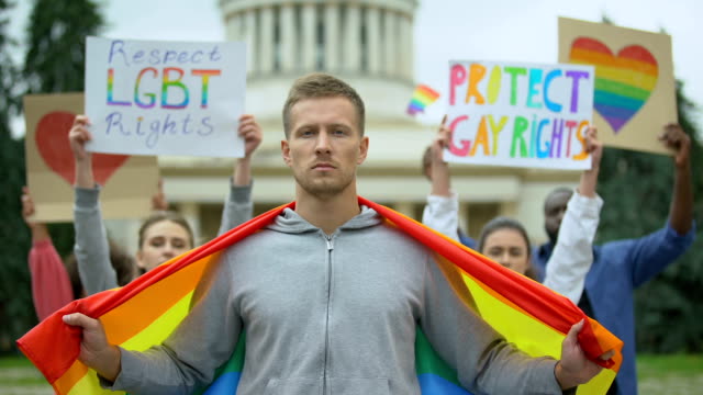 Hombre-guapo-con-bandera-arco-iris-en-medio-de-manifestantes-por-los-derechos-de-los-homosexuales,-evento-de-orgullo-LGBT
