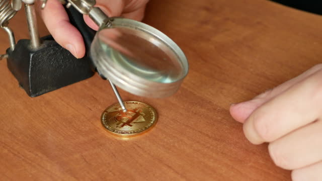 Nahaufnahmen-von-Mann-machen-gefälschte-goldene-Bitcoin-Münze-auf-einem-Schreibtisch-mit-Lupe.-4k-Video-von-Fälscher