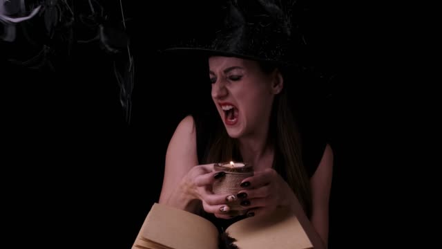 Attractive-witch,-on-a-black-background,-holds-a-candle-over-a-spell-book-in-her-hands-and-screams.-Front-view