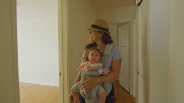 Glückliche-Frau-mit-Baby-in-Träger-zu-Fuß-um-eine-leere-Wohnung