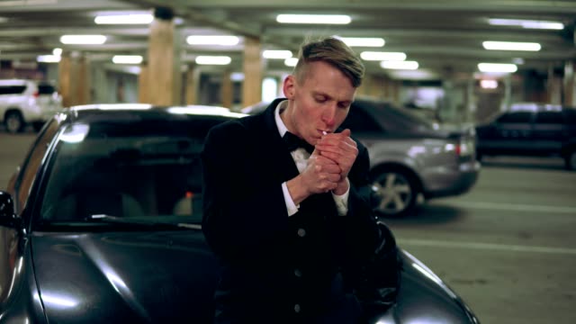 Young-man-in-a-black-suit-with-a-bow-tie-smoking-a-cigarette-sitting-at-the-bonnet-of-the-black-car-in-the-parking.-Waiting-for-someone-and-holding-a-big-bag.-Getting-up-and-going-away.-Front-view