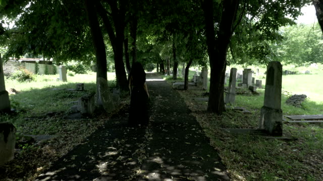 Funeral-gothic-widow-woman-in-black-holding-a-crown-in-hand-walking-in-the-old-cemetery-alley-filled-with-fallen-tree-leaves