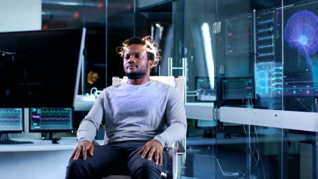 Hombre-usando-ondas-cerebrales-exploración-auricular-se-sienta-en-una-silla-mientras-viendo-estimulantes-imágenes-en-pantalla.-En-el-cerebro-moderno-estudio-laboratorio-monitores-Mostrar-EEG-lectura-y-modelo-de-cerebro.