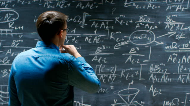 Brilliant-Young-Mathematician-Approaches-Big-Blackboard-and-Finishes-writing-Formula,-Turns-Around-and-Smiles-on-Camera.
