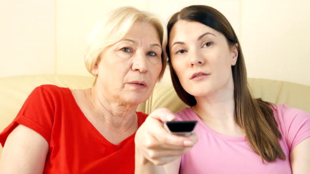 Mother-and-daughter-relaxing-on-sofa-at-home-watching-TV.-Switching-channels-with-remote-control