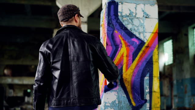 Rear-view-of-male-graffiti-artist-in-leather-jacket-painting-on-damaged-column-inside-empty-industrial-building.-Young-people,-creativity,-casual-clothing-and-modern-art-concept.