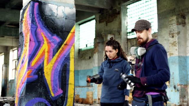 Professional-graffiti-artist-is-teaching-pretty-girl-to-paint-with-aerosol-spray-paint-standing-together-inside-adandoned-warehouse-near-old-column-and-talking.