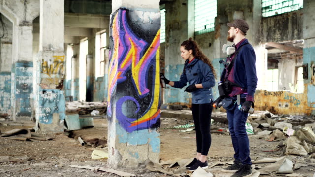Urban-painter-is-explaining-to-attractive-young-woman-how-to-paint-graffiti,-they-are-standing-beside-pillar-in-damaged-empty-warehouse-holding-spray-paint-and-talking.