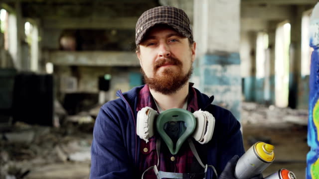 Close-up-portrait-of-handsome-bearded-man-graffiti-artist-standing-inside-abandoned-building-wearing-cap,-gloves-and-pespirator-and-holding-spray-paint