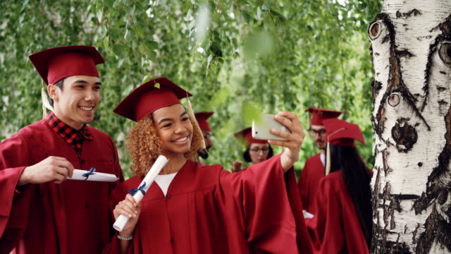 Happy-young-people-girl-and-guy-are-taking-selfie-after-graduation-ceremony-holding-diplomas-wearing-gowns-and-mortarboards.-Photographs-and-education-concept.