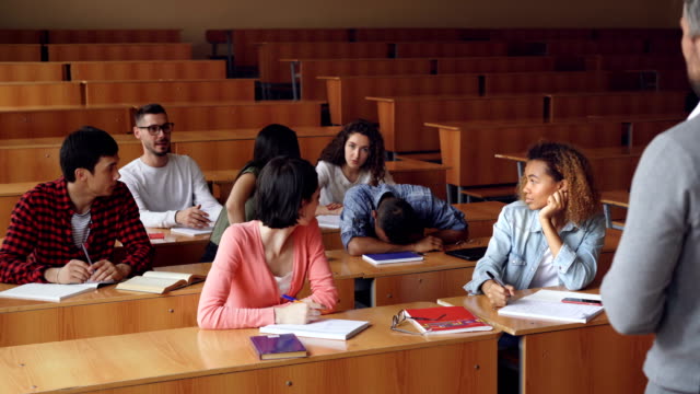 Tired-mixed-race-student-is-sleeping-during-lecture-at-college,-his-classmates-are-waking-him-up,-he-is-standing-and-talking-to-teacher,-young-people-are-laughing.