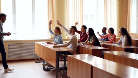 Jóvenes-estudiantes-son-levantando-manos-y-respondiendo-a-las-preguntas-del-profesor-y-barbudo-profesor-de-hombre-es-hablar-y-gesticular.-Ambiente,-luz-de-la-habitación.
