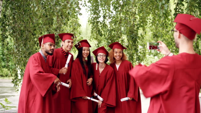 Multiethnic-group-of-graduating-students-is-posing-for-smartphone-camera,-waving-hands-with-diplomas-and-shouting-while-guy-is-shooting-them-touching-screen.