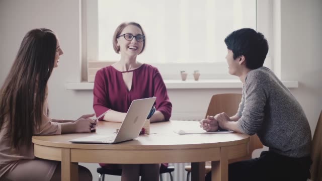 Zwei-schöne-positive-Kaukasische-Mädchen-sprechen-Sie-mit-japanischen-Mann-bei-einem-Vorstellungsgespräch-am-Tisch-in-moderne-gesunde-Büro