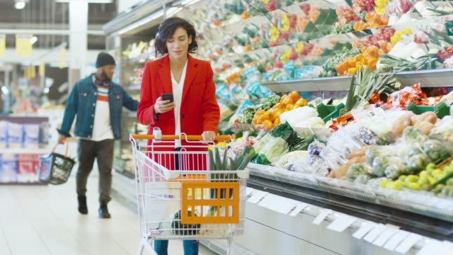 At-the-Supermarket:-Beautiful-Young-Woman-with-Shopping-Cart-Uses-Smartphone-and-Walks-Through-Fresh-Produce-Section-of-the-Store.