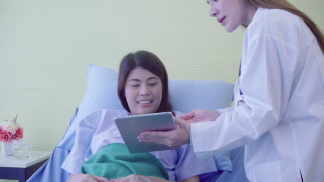 Beautiful-smart-Asian-doctor-and-patient-discussing-and-explaining-something-with-tablet-in-doctor-hands-while-staying-on-Patient's-bed-at-hospital.-Medicine-and-health-care-concept.