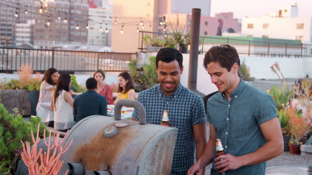 Zwei-Männer-kochen-Barbecue-für-Freunde-versammelten-sich-auf-der-Dachterrasse-mit-Skyline-der-Stadt-im-Hintergrund