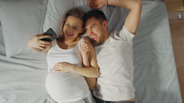 Amantes-de-la-joven-pareja-pasar-mañana-en-la-cama,-mujer-joven-embarazada-muestra-a-su-pareja-algo-en-un-Smartphone-de-pantalla-táctil,-Selfie-de-tomar-y-compartir-imágenes-en-redes-sociales.