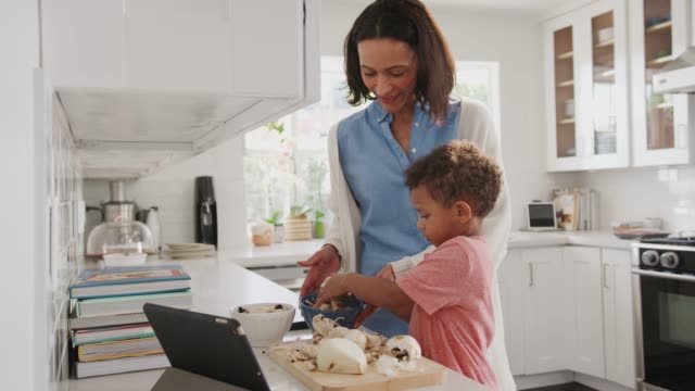Millennial-mother-and-toddler-son-preparing-food-together-in-the-kitchen,-side-view