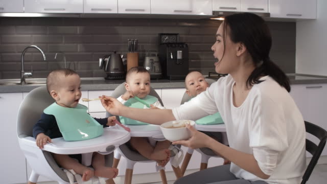 Asiática-mamá-alimentando-el-puré-a-los-trillizos-descuidados-de-1-año-de-edad