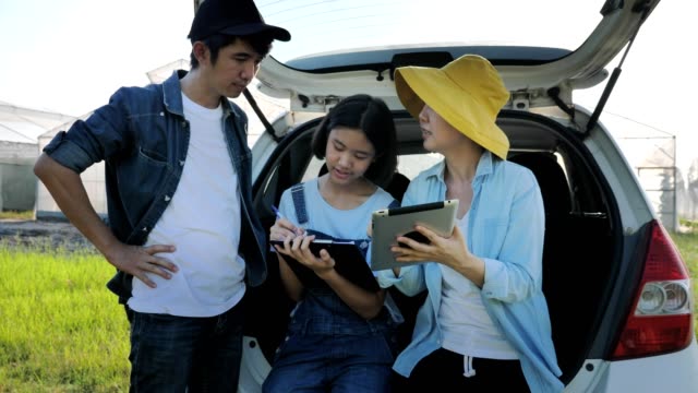 Smart-Asian-family-sitting-on-car-and-using-digital-tablet-for-internet-access-front-of-green-house,-Smart-agriculture-concept