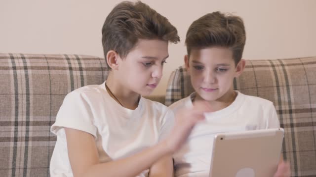 Close-up-faces-of-Caucasian-twin-brothers-playing-video-games-at-tablet-and-giving-high-five.-Siblings-having-fun-together-indoors.-Twins-resting-at-home.