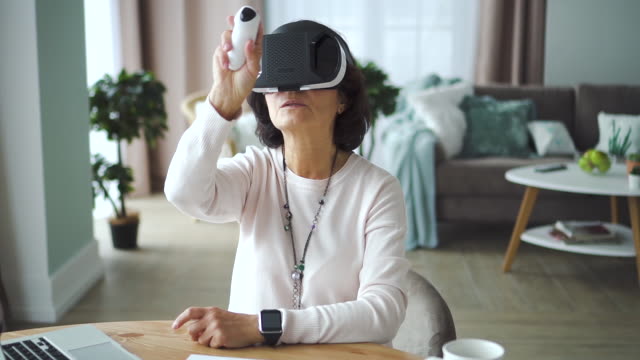 Old-american-woman-with-vr-glasses-sitting-front-laptop-at-table-in-home-room