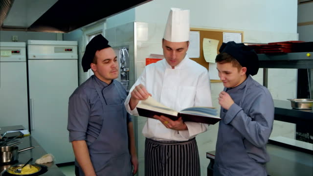 Young-cook-trainees-having-positive-discussion-with-chef-holding-cookbook