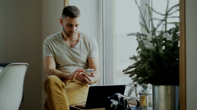 Attractive-young-man-browsing-smartphone-phone-sitting-on-windowsill-with-laptop-and-camera-in-cafe