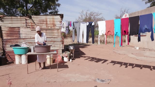 African-woman-without-running-water-doing-laundry-in-a-bucket-in-front-of-her-tin-shack-home