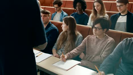 Kamera-mit-Blick-auf-Klasse:-Rückansicht-der-Universität-Professor-gibt-Vorlesung-zu-einem-Klassenraum-voller-Multi-ethnischen-Studenten.-Dozent-Gesticulates-mit-seinen-Händen-während-Kursteilnehmer-hören.