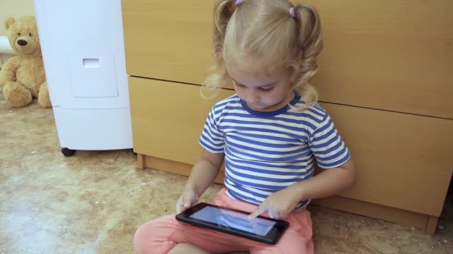 Baby-Mädchen-hält-eine-Tablette-und-sitzt-auf-dem-Boden-im-Zimmer.