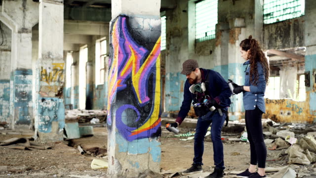 Experienced-graffiti-artist-is-teaching-his-friend-to-create-beautiful-images-with-spray-paint,-they-are-standing-together-in-adandoned-building-near-old-column-and-talking.