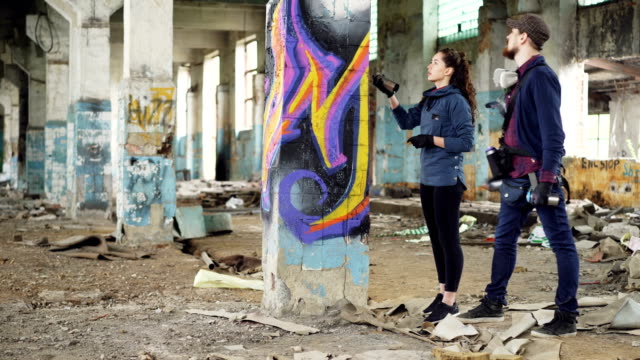 Hübscher-bärtigen-Mann-Graffiti-Künstler-lehrt-seine-Freundin-mit-Sprühfarbe-stehen-in-alten-beschädigten-Gebäude-zeichnen-und-Reden-halten-Spraydosen.