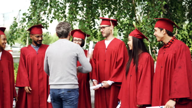 Bärtiger-Mann-stolz-Lehrer-ist-gratuliert-Absolventen-Händeschütteln-und-umarmt-sie-im-Freien-im-College-Campus,-während-Studenten-sprechen-und-hält-Diplome.