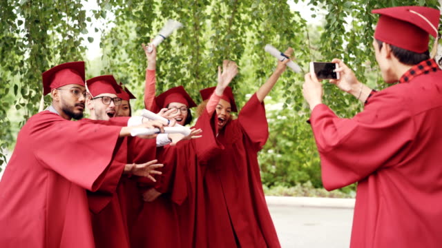 Young-man-with-smartphone-is-taking-pictures-of-graduates-having-fun-posing-with-diplomas-moving-hands-and-fingers-and-shouting-with-happiness.-Education-and-youth-concept.