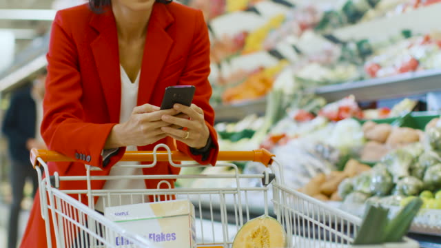 At-the-Supermarket:-Woman-Uses-Smartphone,-Leans-on-Shopping-Cart-in-the-Fresh-Produce-Section-of-the-Store.-In-the-Big-Mall-Woman-Browsing-In-Internet-on-Her-Mobile-Phone.-Slow-Motion.