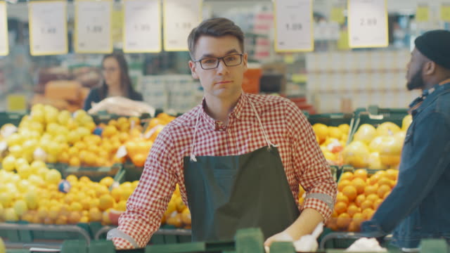 At-the-Supermarket:-Portrait-Of-the-Handsome-Stock-Clerk-Wearing-Apron,-Arranging-Organic-Fruits-and-Vegetables,-He-Smiles-and-Crosses-Arms.-Friendly,-Efficient-Worker-at-the-Store.