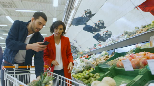 Im-Supermarkt:-glückliche-Paar-macht-Einkaufen,-Auswahl-von-Obst-und-Gemüse-an-der-frischen-Abschnitt.-Mann-nutzt-Smartphone-und-schiebt,-Warenkorb,-Frau-legt-Produkte-in-Wagen.