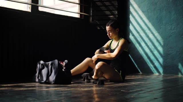 Panning-young-beautiful-woman-sitting-on-floor-and-wrapping-hands-with-black-boxing-wraps-in-club.