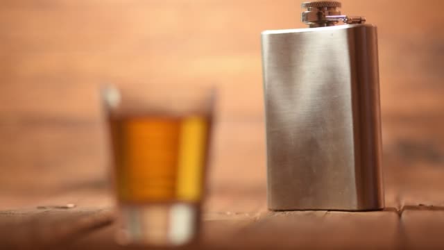 Focus-pull-of-a-whiskey-shot-glass-to-a-metal-hip-flask-in-the-background